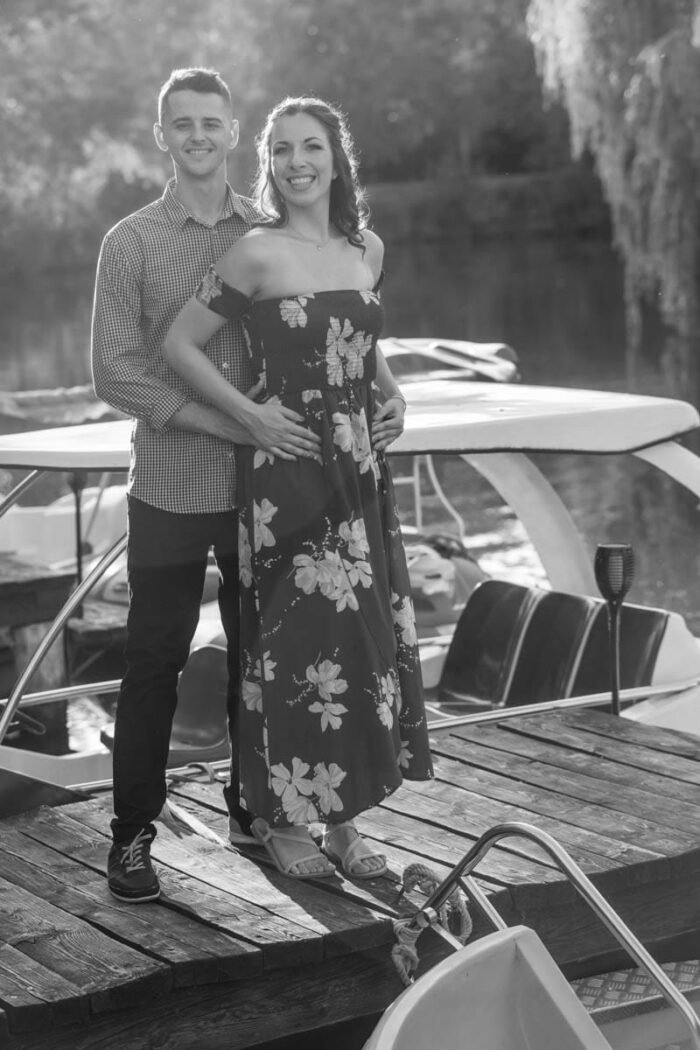 Egy fiatal jegyespár áll egy stégen, háttérben csónakokkal és egy csendes tóval. A férfi szorosan átkarolja a nőt, aki mosolyogva néz a kamerába. A kép fekete-fehér, a napfény sziluetté formálja a párt, fokozva a romantikus hangulatot.