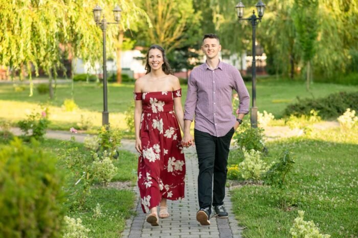 Egy fiatal jegyespár kéz a kézben sétál egy park kanyargós ösvényén, lámpaoszlopok és virágok között. A nő hosszú, virágmintás bordó ruhát visel, a férfi pedig kockás inget és sötét nadrágot. A napsütés és a zöld környezet barátságos, romantikus hangulatot kölcsönöz a képnek. Egy fiatal jegyespár kéz a kézben sétál egy park kanyargós ösvényén, lámpaoszlopok és virágok között. A nő hosszú, virágmintás bordó ruhát visel, a férfi pedig kockás inget és sötét nadrágot. A napsütés és a zöld környezet barátságos, romantikus hangulatot kölcsönöz a képnek. Egy fiatal jegyespár kéz a kézben sétál egy park kanyargós ösvényén, lámpaoszlopok és virágok között. A nő hosszú, virágmintás bordó ruhát visel, a férfi pedig kockás inget és sötét nadrágot. A napsütés és a zöld környezet barátságos, romantikus hangulatot kölcsönöz a képnek.