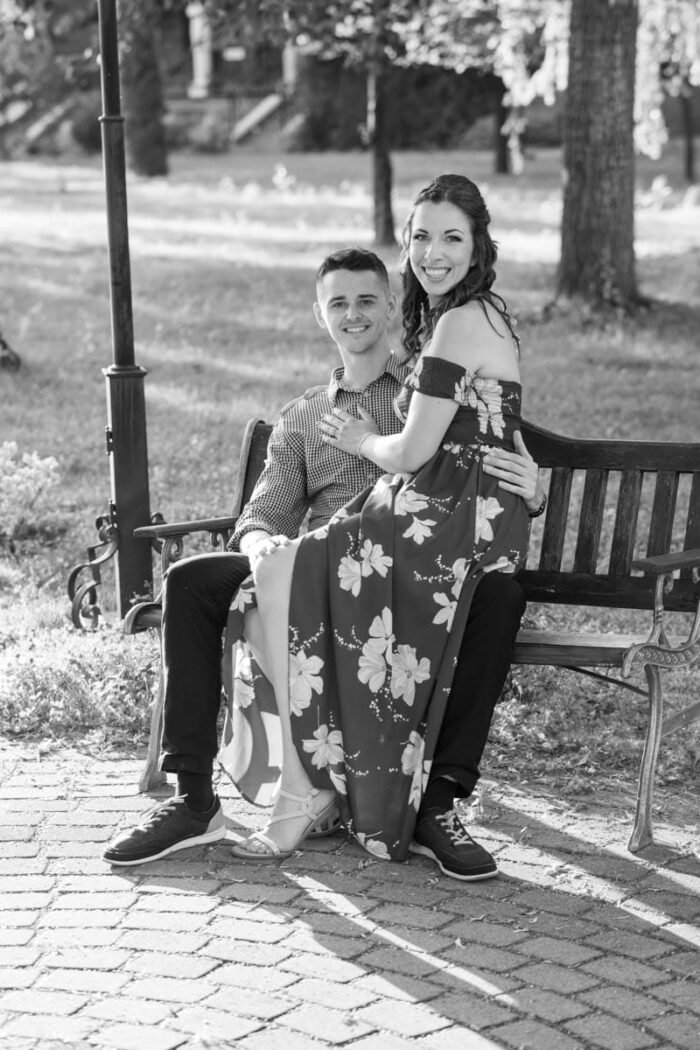 Egy fiatal jegyespár ül egy park padján, a nő a férfi ölében, miközben mosolyognak. A fekete-fehér kép klasszikus és időtlen hangulatot áraszt, a háttérben zöldellő fák és napsütötte park látható.