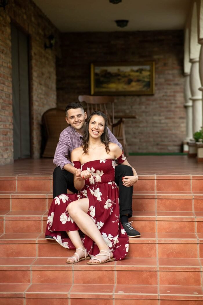 Egy mosolygó jegyespár ül együtt egy verandán lépcsőn, meghitt és otthonos környezetben, miközben egymás kezét fogják. A háttérben rusztikus téglafal és dekoratív bútorok láthatók.