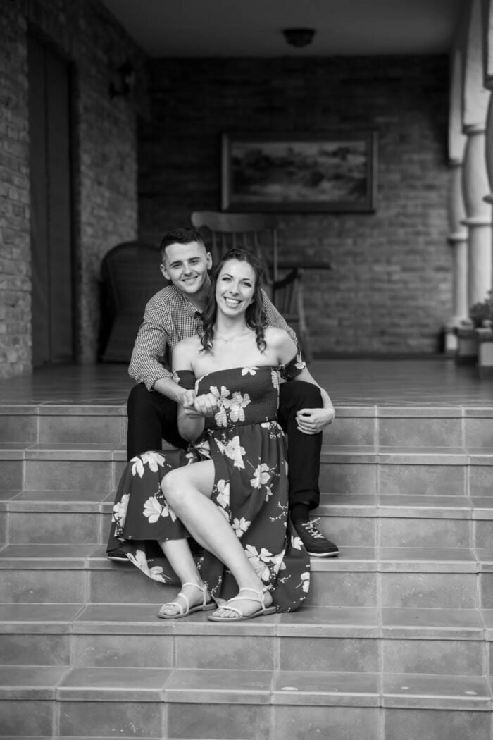 Egy fiatal pár ül együtt egy verandán lépcsőn, egymásra mosolyogva. A fekete-fehér kép időtlen és meghitt hangulatot áraszt, a háttérben otthonos, rusztikus környezettel.