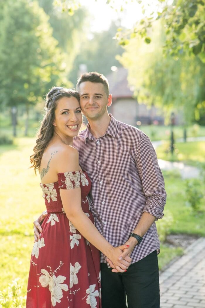 Egy fiatal pár mosolyogva áll egymás mellett egy zöldellő parkban, kézen fogva, a háttérben napfényes természet és egy romantikus épület látható.