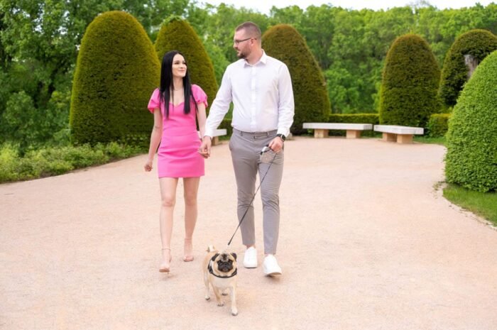Egy jegyespár séta közben kézen fogva, miközben mopsz kutyájukat pórázon vezetik. A nő rózsaszín ruhát és magas sarkút, a férfi fehér inget és világos nadrágot visel. A háttérben gondozott sövények és zöld növényzet teszik idilli hangulatúvá a jelenetet.