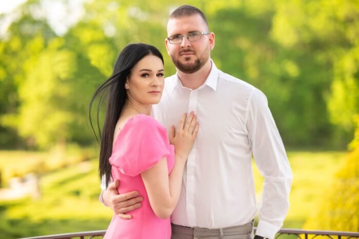 Egy jegyespár áll egymás mellett; a nő rózsaszín ruhában gyengéden a férfi mellkasára helyezi a kezét, míg a férfi fehér inget és világos nadrágot visel. A háttérben buja zöld növényzet és természetes környezet látható.