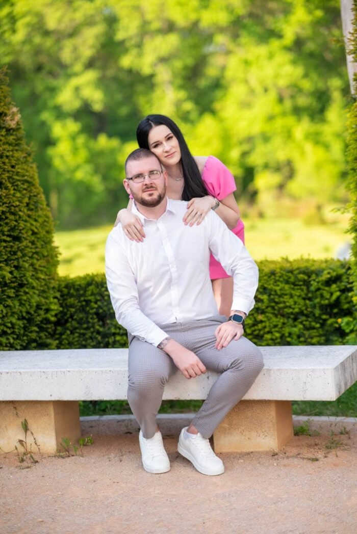 Egy jegyespár ül egy kerti padon; a férfi fehér inget és szürke nadrágot visel, míg a nő rózsaszín ruhában áll mögötte, gyengéden a vállára támaszkodva. A háttérben buja zöld növényzet és fák teremtik a romantikus hangulatot.