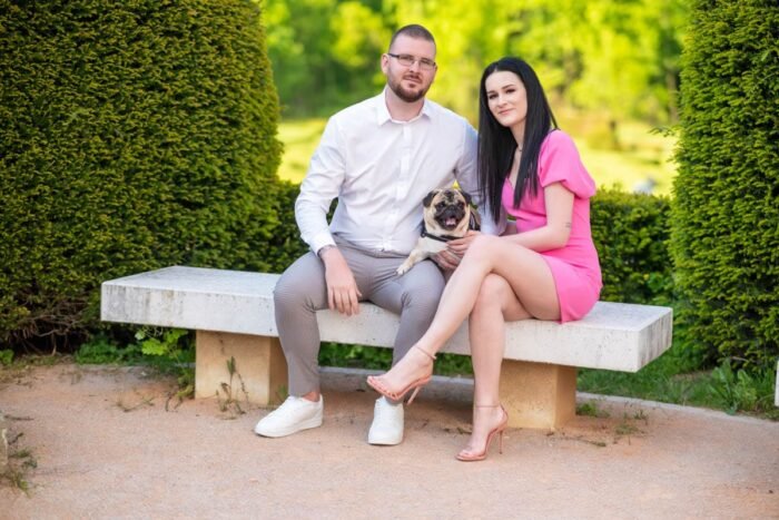 Egy pár ül egy kerti padon, miközben középen a mopsz kutyájuk ül mosolyogva. A nő rózsaszín ruhát és magas sarkút, a férfi fehér inget és világos nadrágot visel. A háttérben buja zöld növényzet és fák teszik hangulatossá a képet.