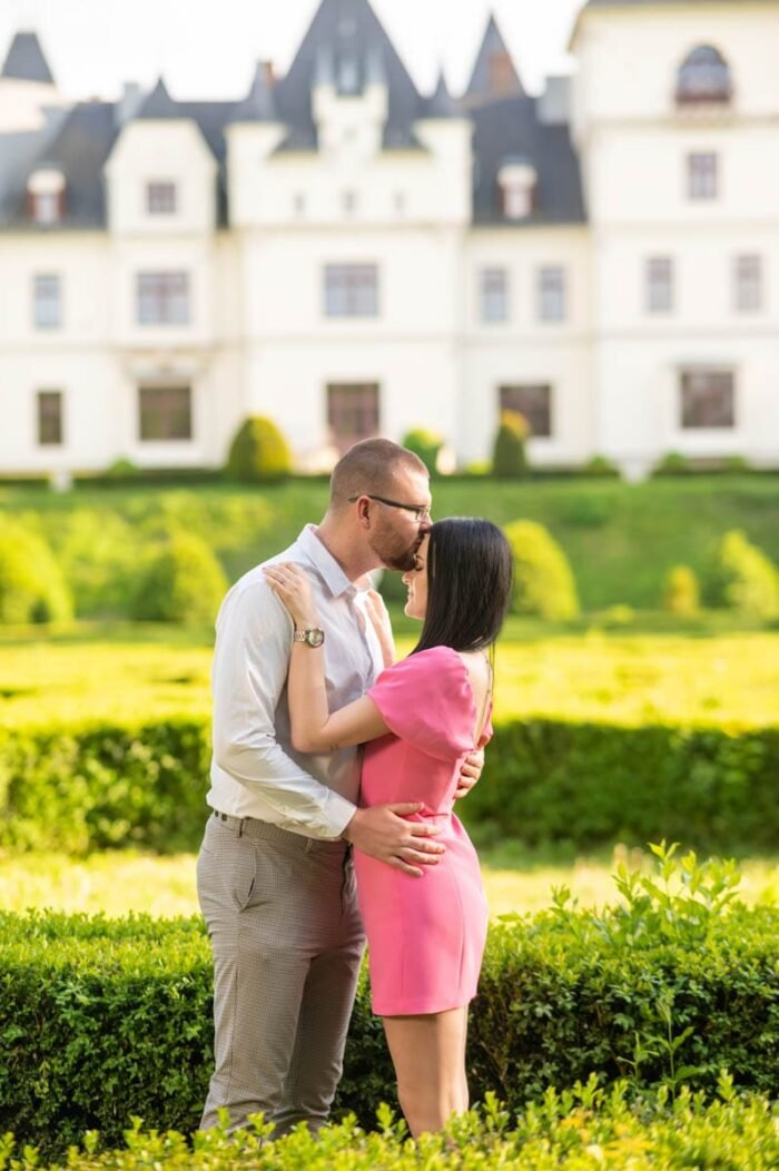 Egy pár csókot vált egy gyönyörű kastély kertjében; a nő rózsaszín ruhát visel, a férfi fehér inget és világos nadrágot. A háttérben gondozott sövények és a kastély épülete látható.