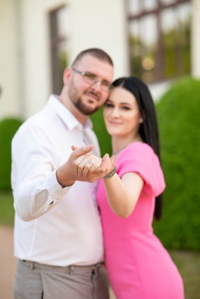 Egy pár kéz a kézben mutatja az eljegyzési gyűrűt, miközben egymásra mosolyognak. A férfi fehér inget, a nő élénk rózsaszín ruhát visel. A háttérben zöld bokrok és egy épület homályosan látszanak.