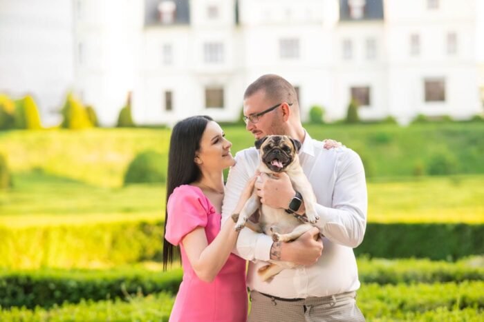 Egy pár a parkban áll, kezükben egy mopsz kutyával. A nő élénk rózsaszín ruhában, a férfi fehér ingben és szemüvegben látható. A háttérben egy elegáns épület és gondozott kert teremt idilli hangulatot.