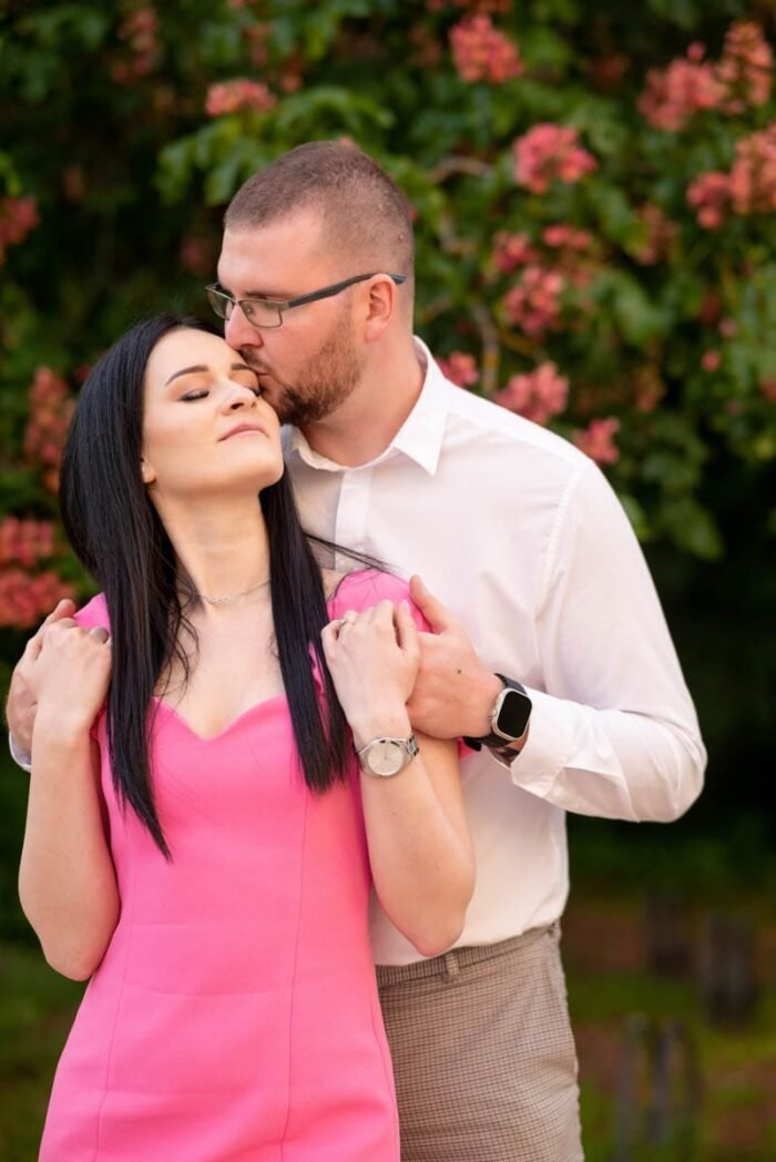 Egy pár romantikus pillanatot oszt meg egy virágzó bokor előtt; a férfi gyengéden megpuszilja a nő homlokát, miközben a nő csukott szemmel élvezi a pillanatot. A nő élénk rózsaszín ruhát, a férfi fehér inget visel.