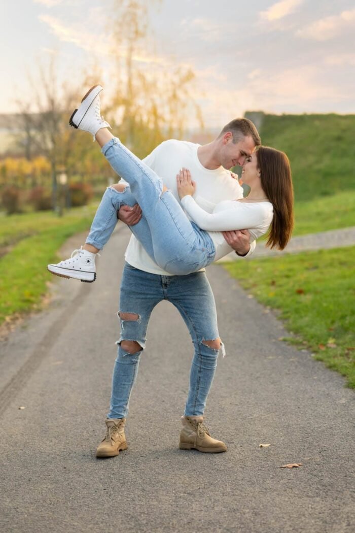 Egy jegyespár romantikus pillanatban, ahol a férfi karjaiban tartja a mosolygó nőt, miközben egymás szemébe néznek. A háttérben őszi táj és lenyugvó nap adja meg a különleges hangulatot.