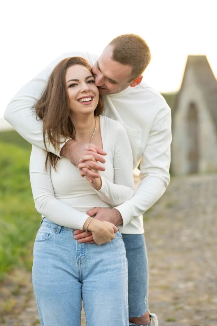 Egy mosolygó fiatal nő és párja, aki gyengéden átöleli és egy csókot lehel a hajára, egy köves úton állnak egy vidéki tájban, harmóniát és boldogságot sugározva.