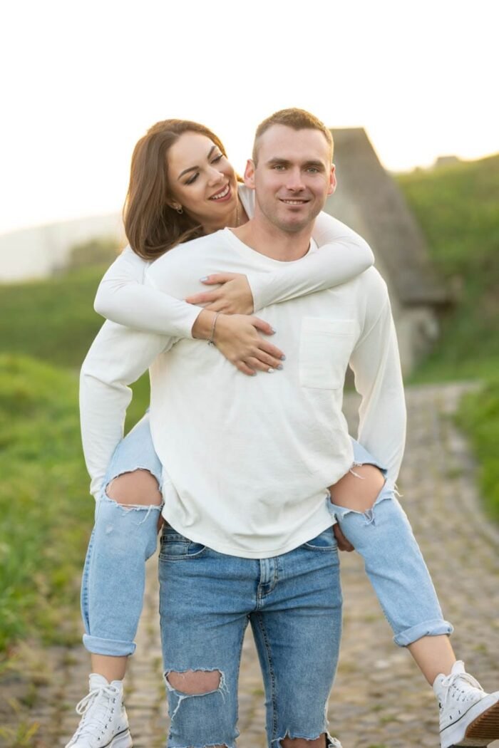 Egy fiatal nő vidáman átöleli párját a hátán ülve, miközben egy köves úton sétálnak egy zöld, vidéki tájon. A mosolygó pár könnyed és boldog hangulatot áraszt.