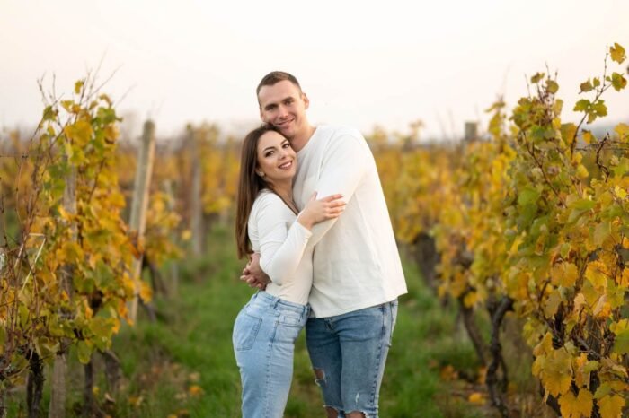 Egy fiatal pár ölelkezve áll egy őszi szőlőültetvény közepén, mosolyogva és boldogan, a háttérben az aranyló őszi levelek és a természet harmóniája.