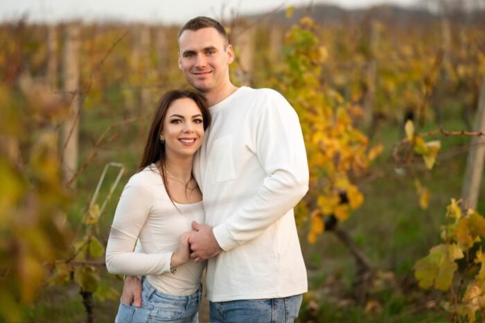 Egy fiatal pár áll egymás mellett egy őszi szőlőültetvényben, mosolyogva, a háttérben az ősz aranyló színeivel, amely meghitt és természetes pillanatot sugároz. Páros fotózás Debrecenben