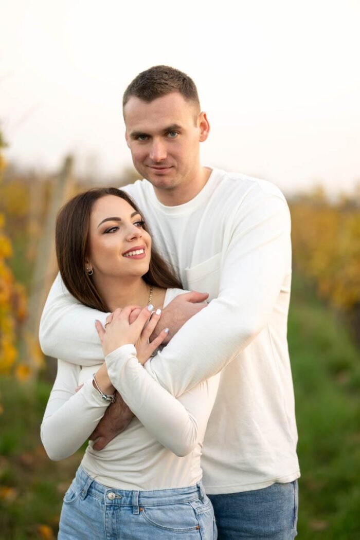 Egy fiatal pár áll egymást átölelve egy őszi szőlőültetvény mellett, mosolyogva, harmóniát és boldogságot sugározva a természetben.