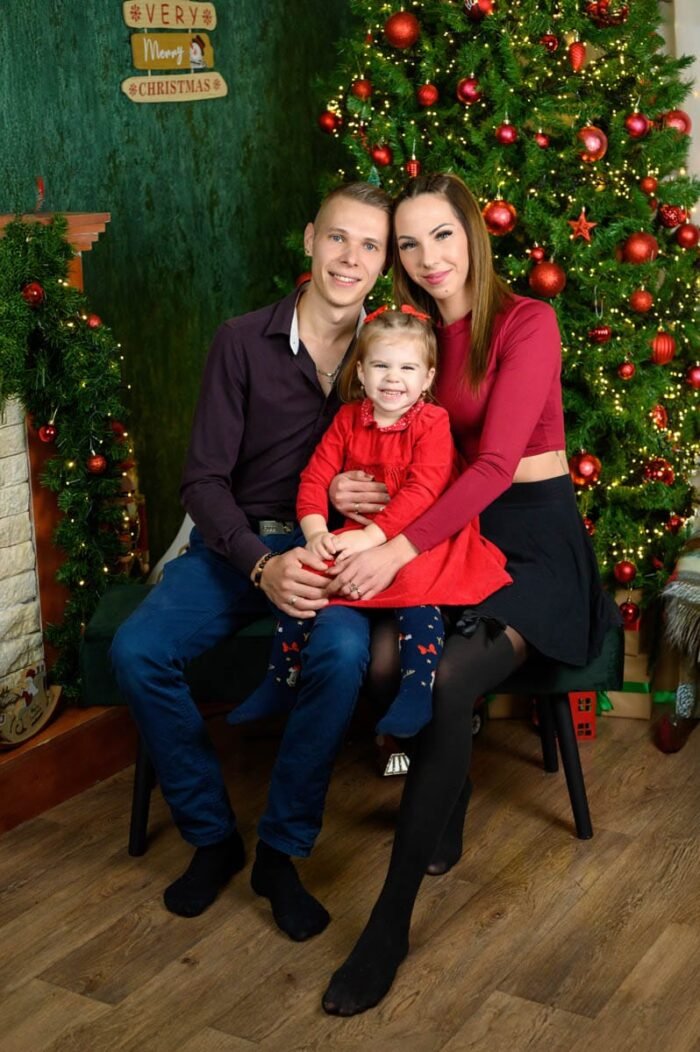 Háromtagú család közös karácsonyi fotója: fiatal szülők kislányukkal egy széken ülnek a feldíszített karácsonyfa előtt, mindannyian mosolyognak.