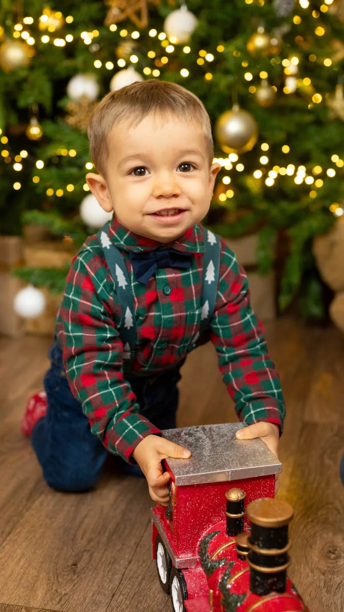 Kisfiú kockás karácsonyi ingben és farmerben egy piros játék mozdonnyal játszik a karácsonyfa előtt. A háttérben arany gömbök és meleg fények díszítik a fát.