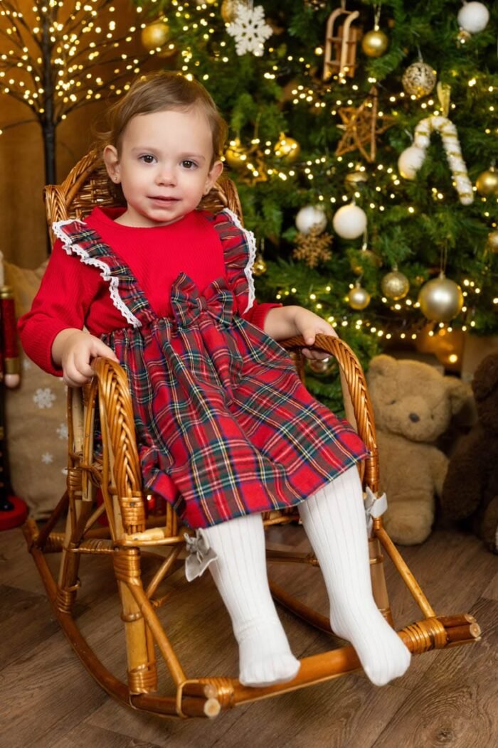 Kislány piros-kockás karácsonyi ruhában, fehér harisnyában ül egy fonott hintaszékben. A háttérben arany és fehér díszekkel, fényekkel feldíszített karácsonyfa látható.