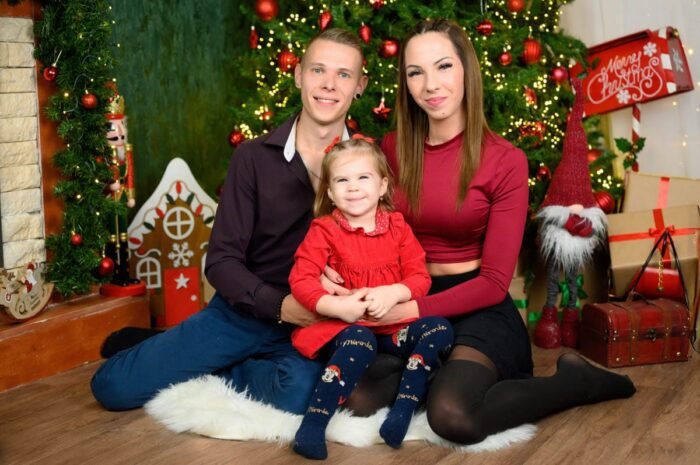 Családi fotózás karácsonyi dekoráció előtt, a szülők és a gyermek boldogan mosolyognak a karácsonyfa mellett