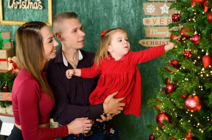 Családi fotózás a karácsonyi dekoráció előtt, a kisgyermek díszít egy karácsonyfát a szülei társaságában