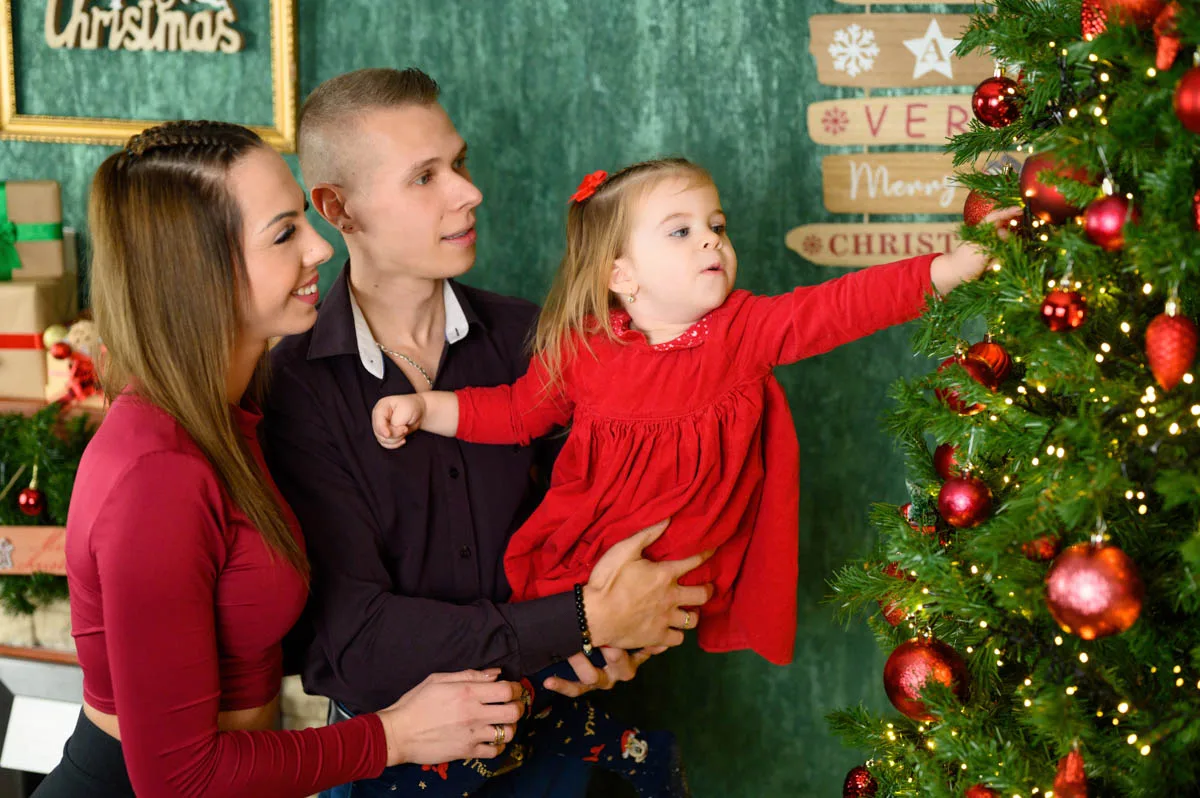 Családi fotózás a karácsonyi dekoráció előtt, a kisgyermek díszít egy karácsonyfát a szülei társaságában