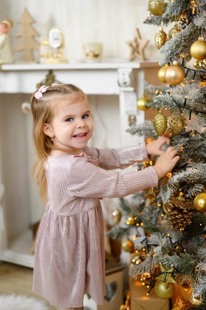 Karácsonyi fotózás Debrecenben. Egy kislány boldogan díszíti a karácsonyfát arany színű díszekkel, miközben mosolyog. A háttérben karácsonyi dekorációk és ajándékok láthatók.