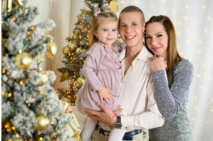 Egy mosolygós család a karácsonyi dekorációk között: egy fiatal pár és kislányuk állnak egymáshoz közel, karácsonyfák és ünnepi fények díszítik a hátteret.