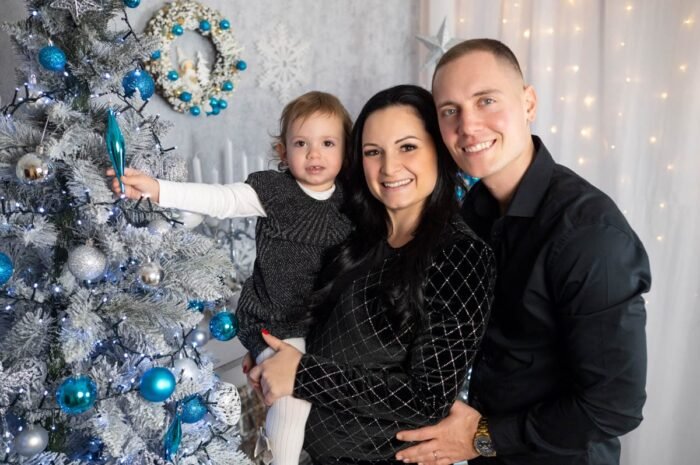 Háromtagú család egy ezüst-kék díszítésű fehér karácsonyfa előtt: kislányuk szürke pulóverben egy kék díszt tart, édesanyja fekete mintás ruhában várandósan, édesapja fekete ingben mosolyog. A háttérben kék gömbök és fényfüzér.