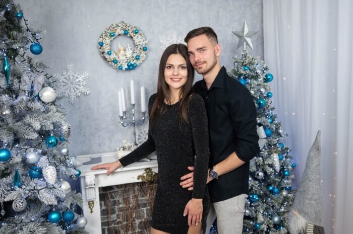 Egy fiatal pár áll egy karácsonyi díszletben: a nő fekete csillogó ruhában, a férfi fekete ingben és világos nadrágban. A háttérben fehér-kék díszítésű karácsonyfák, kandallópárkány és egy kék gömbökkel díszített koszorú látható.