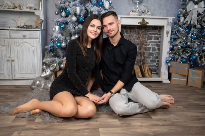 Fiatal pár ül egy szőrmés szőnyegen a karácsonyi díszlet előtt. A nő fekete csillogó ruhában, a férfi fekete ingben és világos nadrágban. Háttérben fehér-kék díszítésű karácsonyfák, kandallópárkány és ajándékdobozok láthatók.
