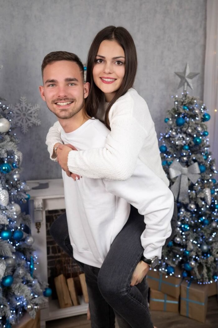 Fiatal pár fehér pulóverben, a férfi a hátán viszi a nőt, mindketten mosolyognak. A háttérben ezüst-kék díszítésű karácsonyfa, kandallópárkány és ajándékdobozok láthatók.