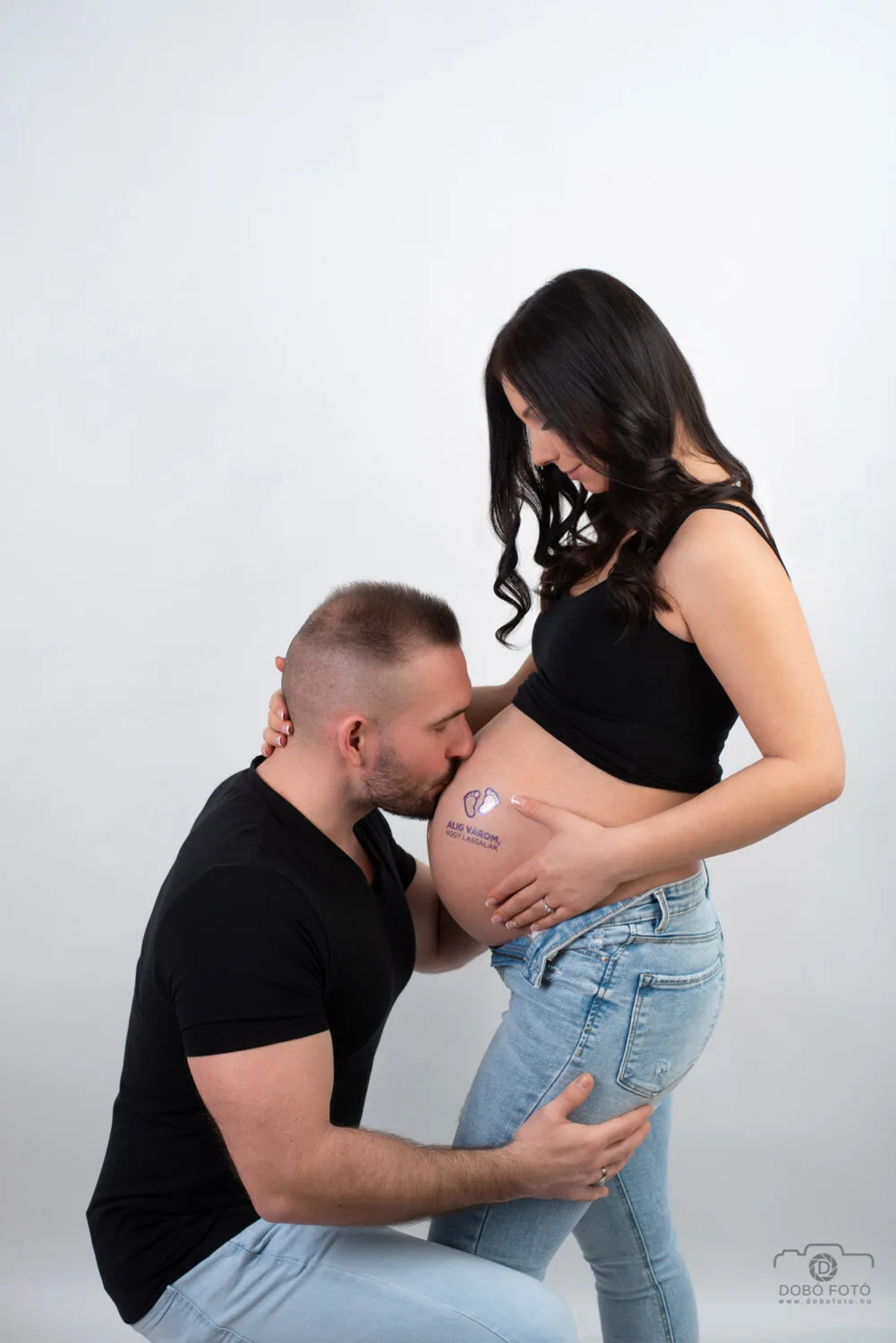 Várandós fotózás Debrecenben - Intim kismama fotó: apa gyengéden megcsókolja a kismama pocakját, fekete pólóban és világos farmerben, baba lábnyom tetoválással.
