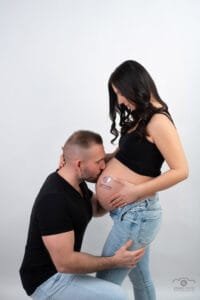 Várandós fotózás Debrecenben - Intim kismama fotó: apa gyengéden megcsókolja a kismama pocakját, fekete pólóban és világos farmerben, baba lábnyom tetoválással.