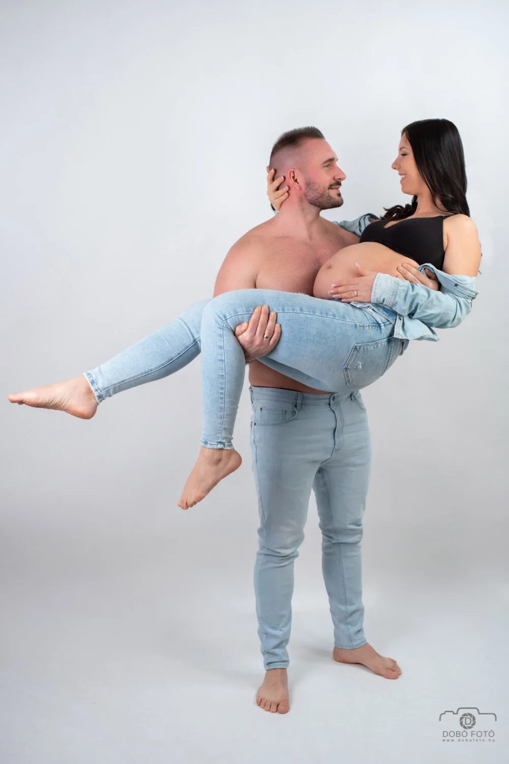 Kismama fotózás Debrecenben - Romantikus kismama páros fotó: apa karjaiban tartja várandós párját, világos farmerben, fekete felsőben, fehér háttér előtt. Dobó Fotó műtermi kismama fotózás.