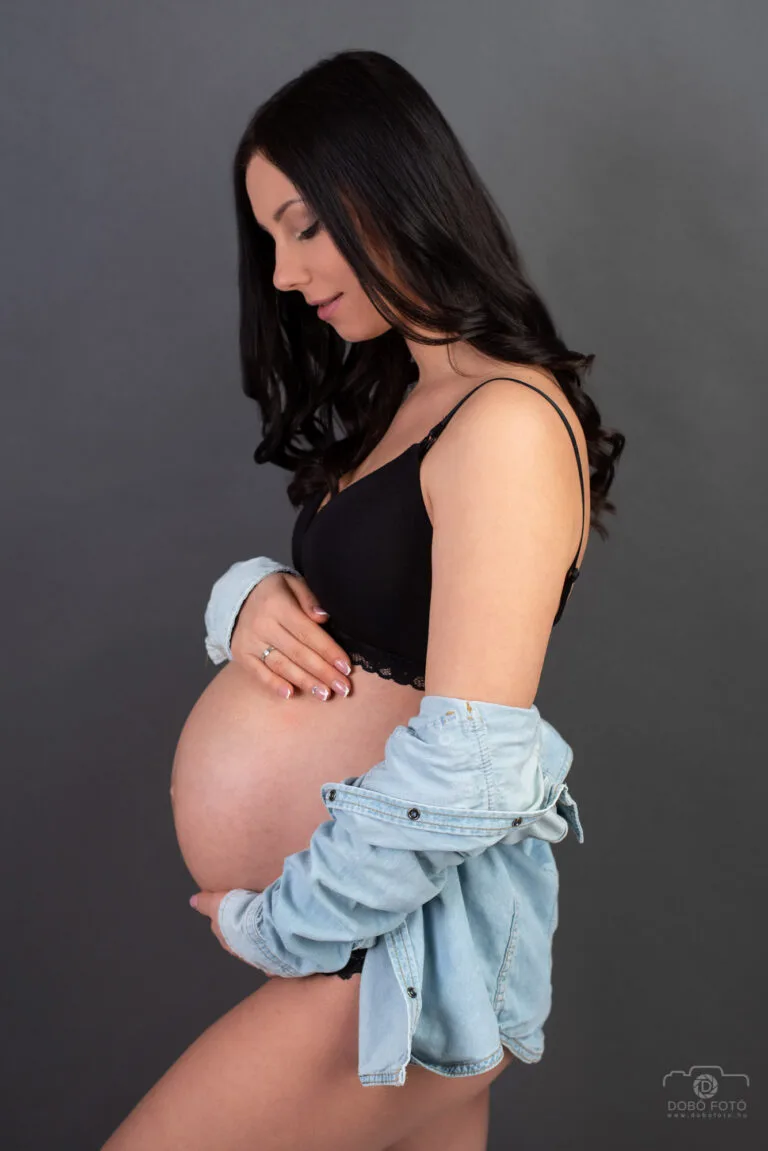 Művészi kismama portré Debrecenben - fekete felsőben és farmerdzsekiben álló várandós nő, profilból fotózva, sötét háttér előtt.
