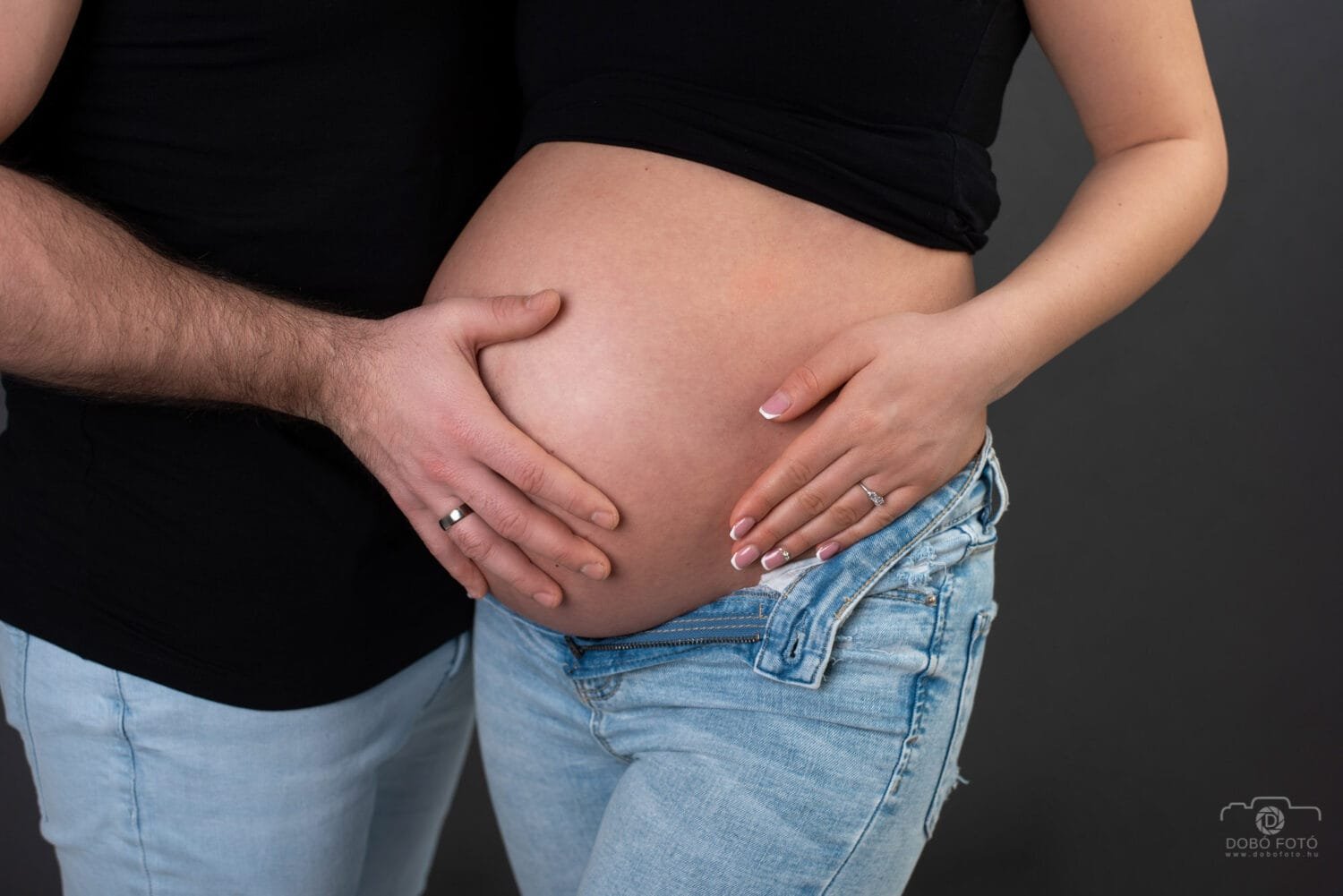 Debreceni kismama fotózás - várandós pocak közelképe, a szülők kezei gyűrűkkel a pocakon, fekete felső és világos farmer viseletben, sötét háttér előtt.