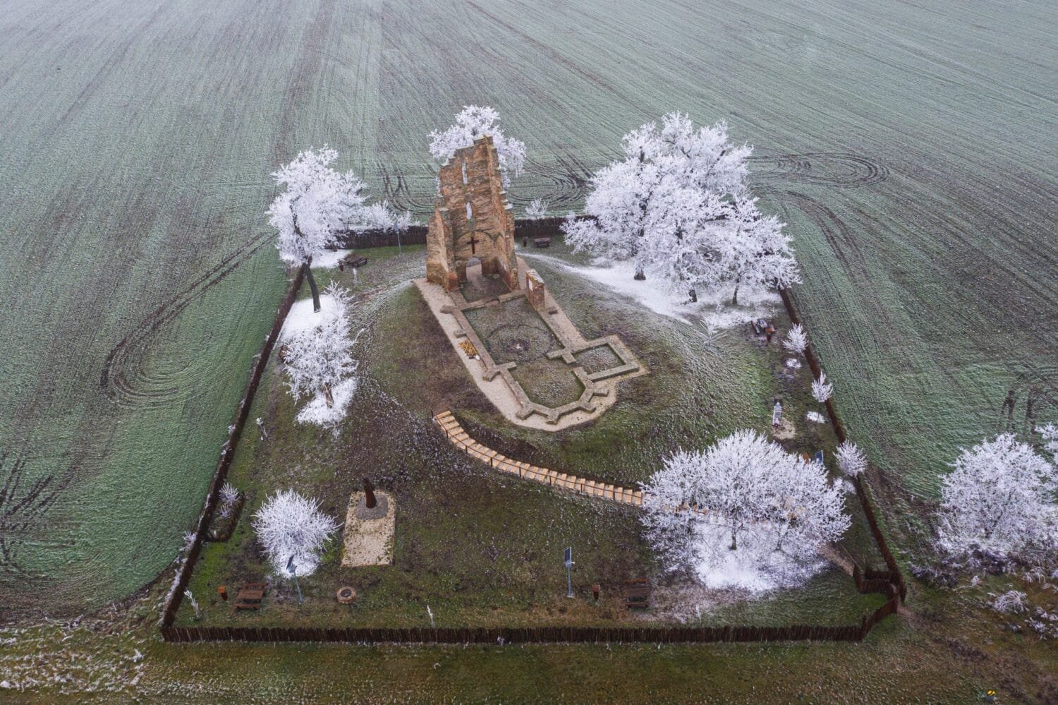 Légifelvétel egy romtemplomról, amelyet hóval borított fák és zöld mező vesz körül. A romtemplom körüli gyalogos ösvények és kerítés is látható, ünnepélyes téli hangulatot teremtve.