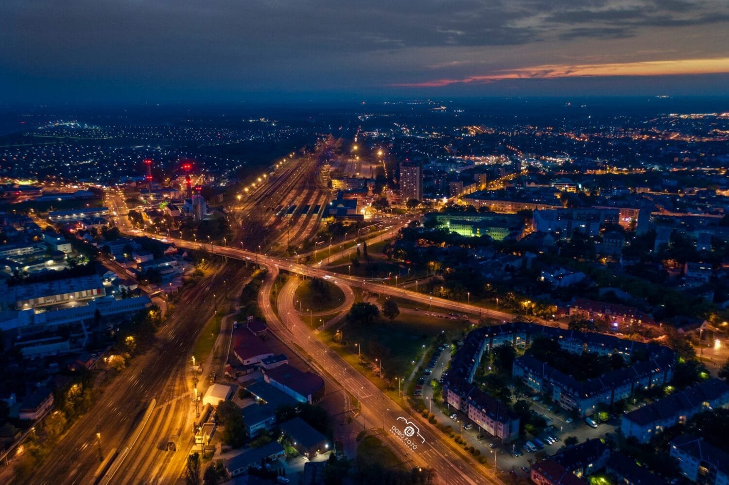 Légifelvétel Debrecen városáról éjszaka, kivilágított utak, vasútvonalak és épületek láthatók, a háttérben a naplemente halvány fényével.