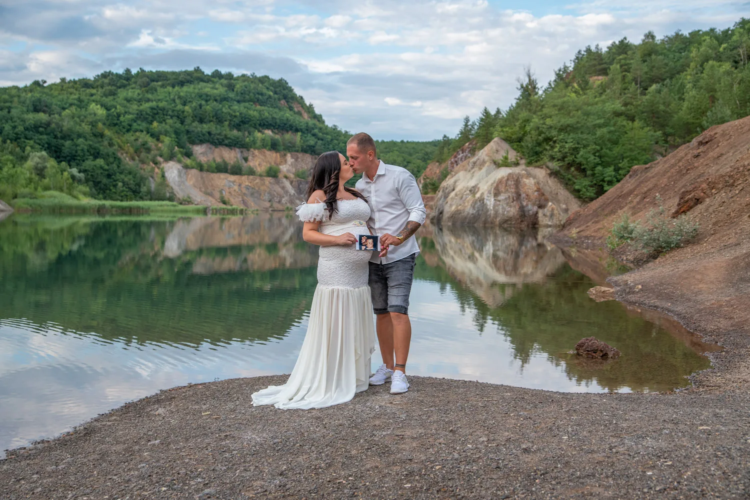 Kismama fotózás hajdúböszörményben - Egy szerelmes pár áll a tó partján, a férfi és a nő egymásba karolva, meghitten csókolóznak. A nő elegáns, fehér kismamaruhát visel, míg a férfi laza fehér ingben és farmer rövidnadrágban van. A férfi kezében egy ultrahangos kép található, amely szimbolikusan kiemeli a családi boldogságot. A háttérben a gyönyörű Rudabányai bányató tükröződő vízfelülete és a zöld növényzet teszi a fotót természetközelivé és varázslatossá.