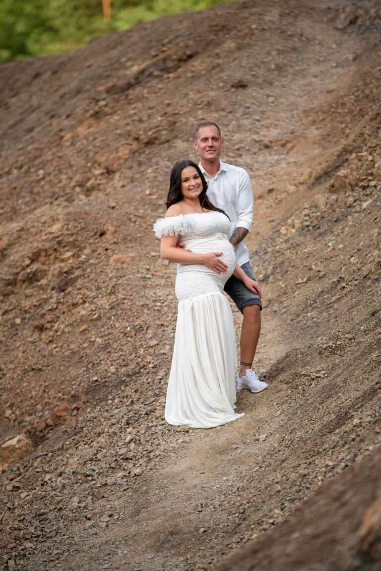 Kismama fotózás Hajdúböszörmény - Egy pár kismama fotózáson, ahol a nő földig érő, hófehér kismamaruhát visel, miközben a férfi szorosan mögötte áll, fehér ingben és farmer rövidnadrágban. A fotó háttere egy meredek, köves hegyoldal, amely természetes, rusztikus hangulatot kölcsönöz a képnek. A nő mosolyog, miközben kezét a pocakjára helyezi, kihangsúlyozva az intim és boldog pillanatot.