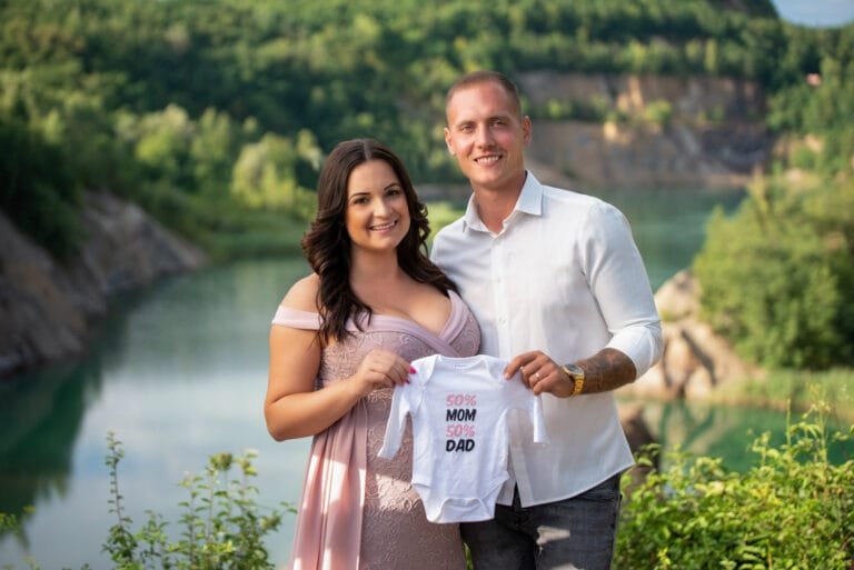 Kismama fotózás Hajdúszobolszó. Várandós pár kismama fotózáson, kezükben egy babaruhával, természetes tóparti háttérrel. Romantikus kismama pillanatok szabadtéren.