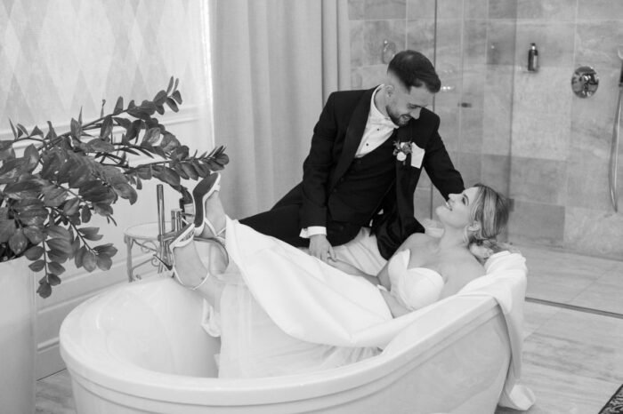 Romantikus és játékos pillanat: a menyasszony a fürdőkádból mosolyogva figyeli a vőlegényt, megörökítve a pillanat különlegességét és a pár közötti kötetlen intimitást.
