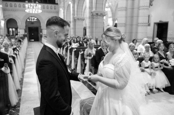 A menyasszony mosolyogva húzza fel a gyűrűt a vőlegénye ujjára, megörökítve a meghitt pillanatot, amely az összetartozásukat szimbolizálja.