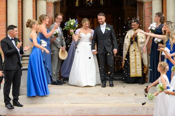 A friss házaspár boldogan lép ki a templom kapuján, miközben a vendégek tapsolva és virágszirmokat szórva ünneplik őket.