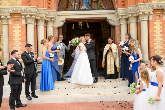 A friss házaspár megcsókolja egymást a templom előtt, miközben a nap fénye lágyan megvilágítja őket, és a vendégek boldogan tapsolnak az emlékezetes pillanatért.