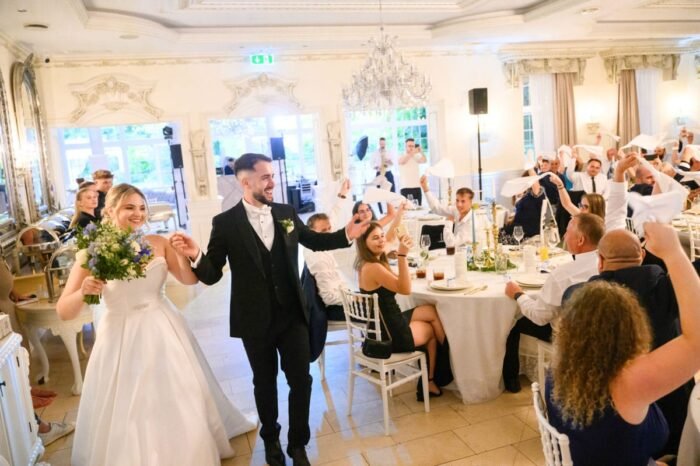 Az ifjú pár kéz a kézben vonul be az ünnepi helyszínre, mosolyogva és a vendégek szeretetteljes tapsától kísérve, miközben a pillanat varázslata mindenkit magával ragad.