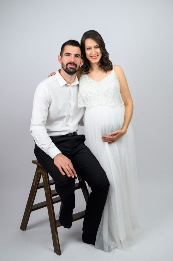 Egy fiatal pár boldogan mosolyog a kamera felé. A kismama kezét szeretetteljesen helyezi a pocakjára, míg párja mellette ül egy fa létrán, karjával gyengéden átölelve őt. A kép a családi boldogság és az új élet érkezésének örömét sugározza.