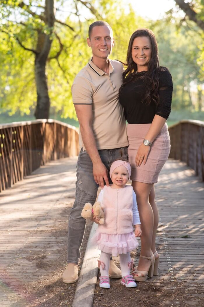 Egy fiatal pár és kislányuk mosolyog egy napsütéses hídon, gyönyörű természetes háttérrel. A kislány plüssállatot tart a kezében, a szülők szeretetteljesen mellette állnak.