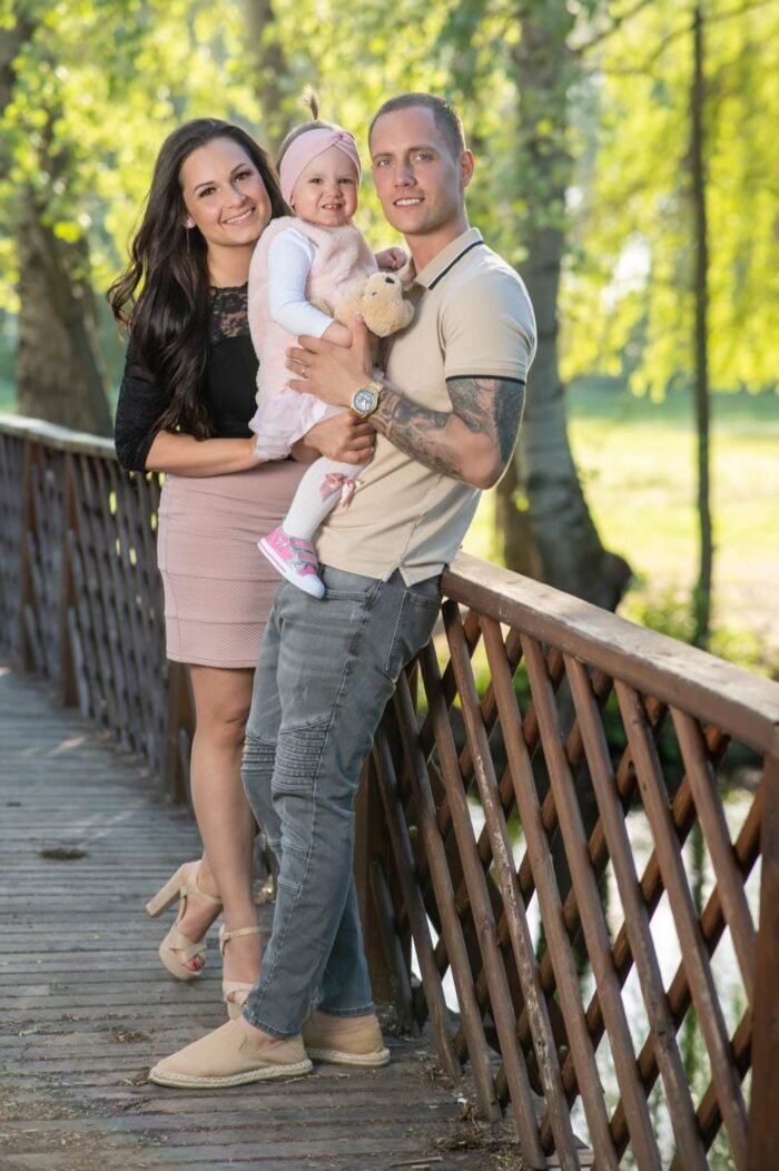 Egy boldog család a hídon állva, a szülők karjaikban tartják kislányukat, aki plüssállatot szorongat. A háttérben zöldellő fák és napsütés teszik idillivé a jelenetet.